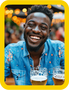 A man with short curly hair smiles while sitting at an outdoor event, wearing a denim shirt with colorful patterns. Two glasses of beer are on the table in front of him, and string lights create a festive atmosphere in the blurred background. The image is framed by a yellow border.