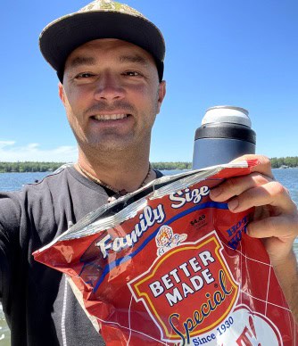 A man wearing a baseball cap smiles while holding a large red bag of Better Made Special Family Size Potato Chips in one hand and a thermos in the other. He stands outdoors with a lake and trees in the background on a sunny day.