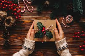 A pair of hands wearing a cozy sweater holds a wrapped holiday gift decorated with holly leaves and berries. The gift is set on a wooden table surrounded by festive items like candy canes, pinecones, red berries, and twine, evoking a holiday atmosphere.